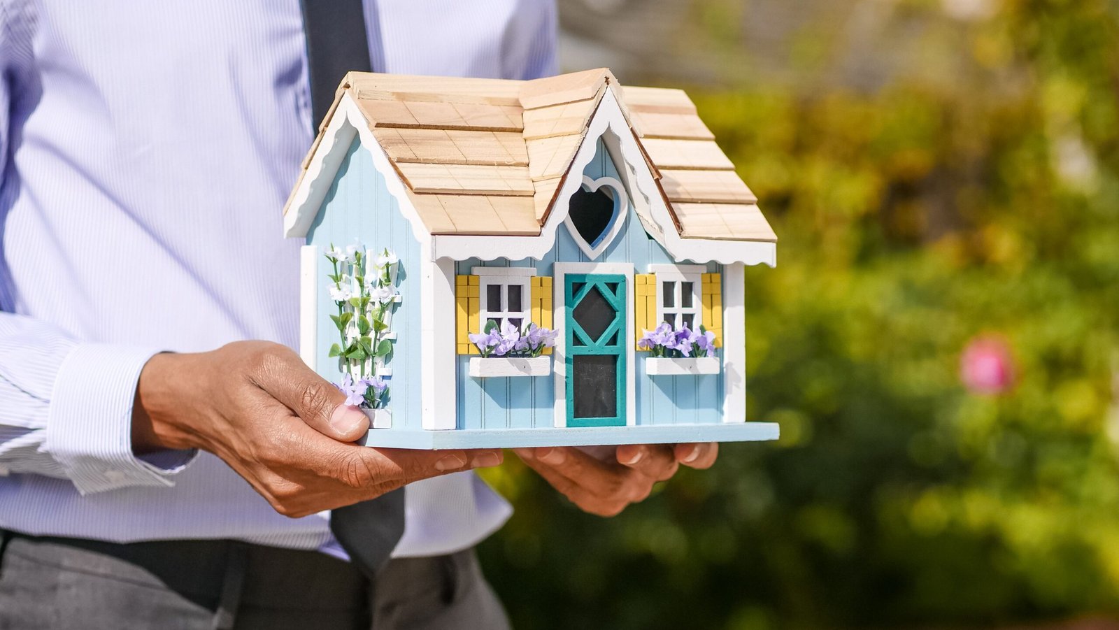 A miniature blue cottage-style birdhouse held in two hands, symbolizing a tiny home